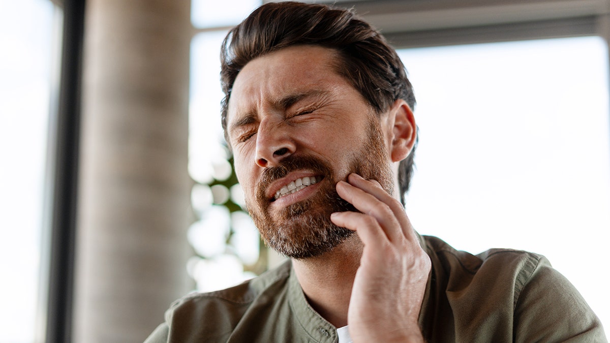 Young man wincing in pain with eyes closed as he touches jaw, indicating toothache.