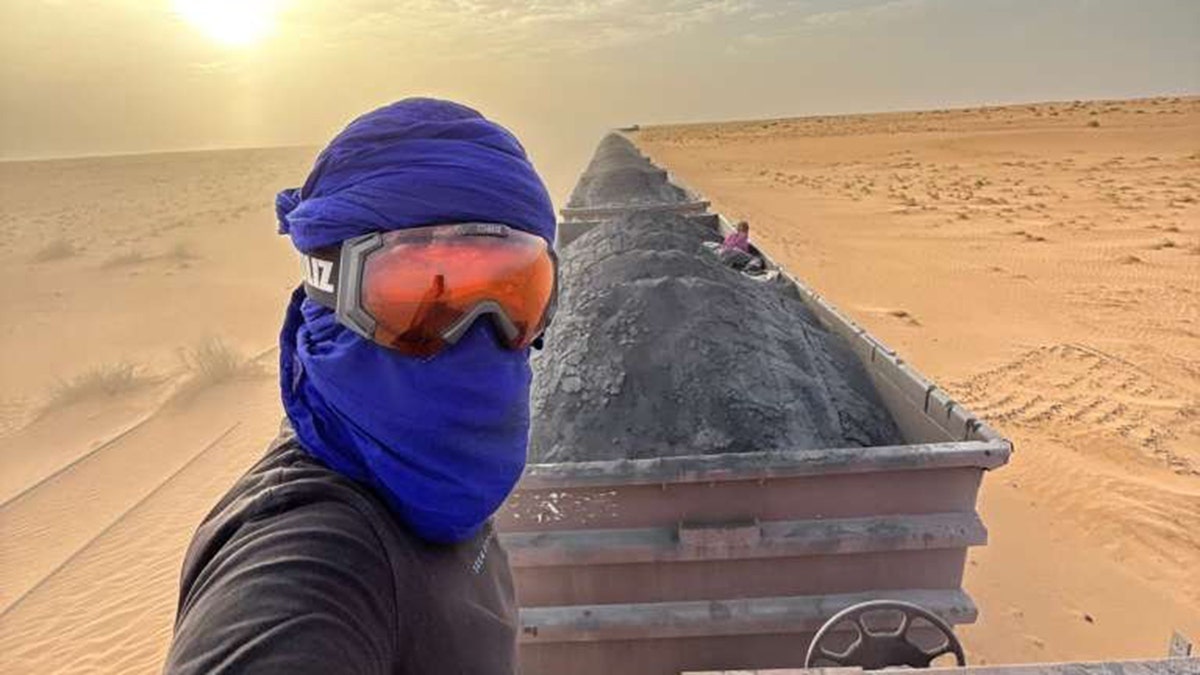 Person wearing blue scarf and goggles rides atop an iron ore train crossing a desert.