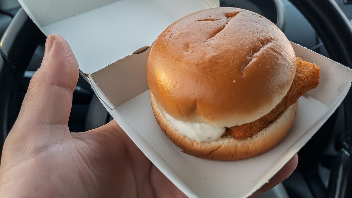 Hand holding McDonald's filet-o-fish sandwich in paper takeout box in car. Tartar sauce seen oozing from one side.