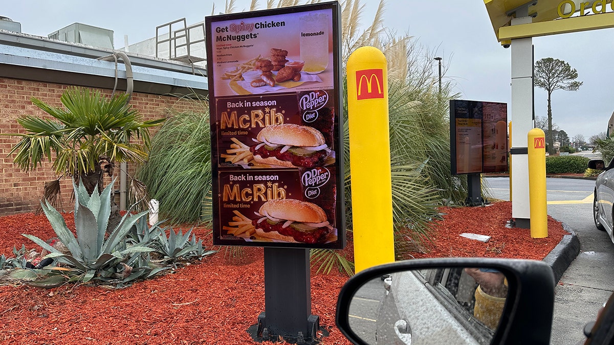 Drive-thru signs seen advertising McDonald's McRib, with person in driver seat taking pic from car, sideview mirror seen at bottom right in drive-thru line.