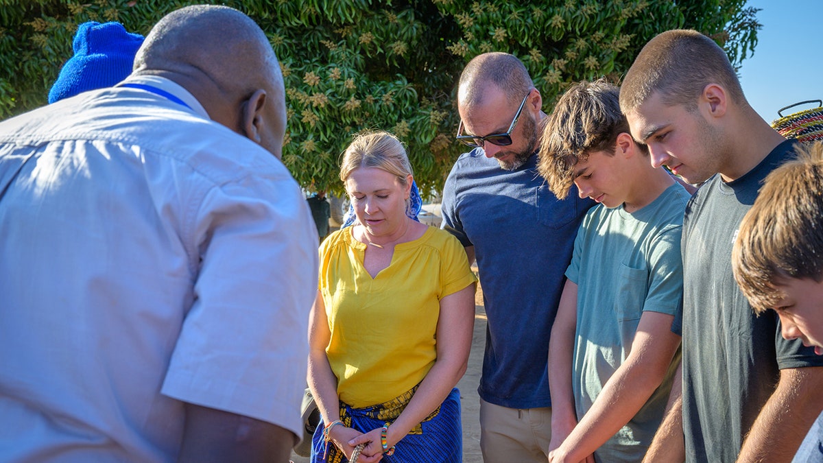 Melissa Joan Hart and a group of people stand in a circle outdoors with their heads bowed, appearing to participate in a moment of reflection or prayer.