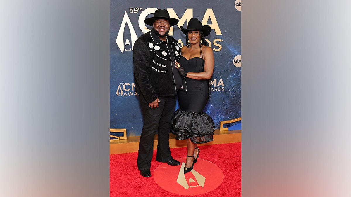 Michael Trotter Jr. and Tanya Trotter of The War and Treaty pose together on the red carpet at the 59th Annual CMA Awards in Nashville, Tennessee, on November 19, 2025. He wears a black jacket with white floral embroidery and a cowboy hat, while she wears a strapless black ruffled dress and matching hat.
