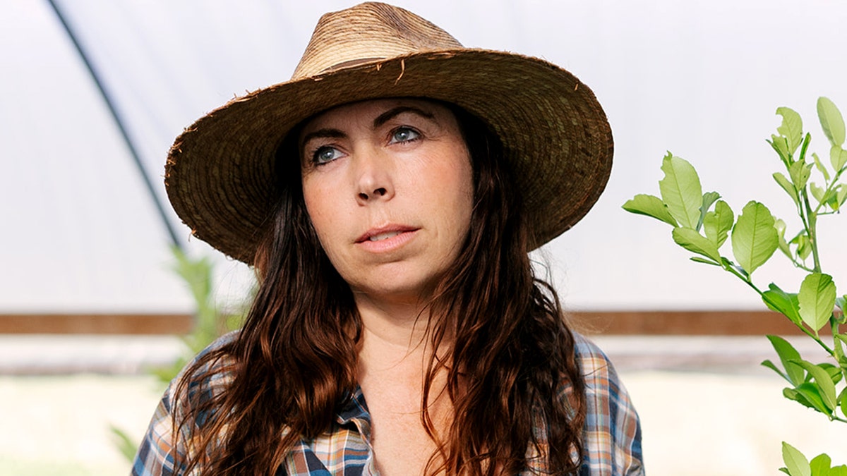 Mollie Engelhart wears a hat and looks up from inside her greenhouse.