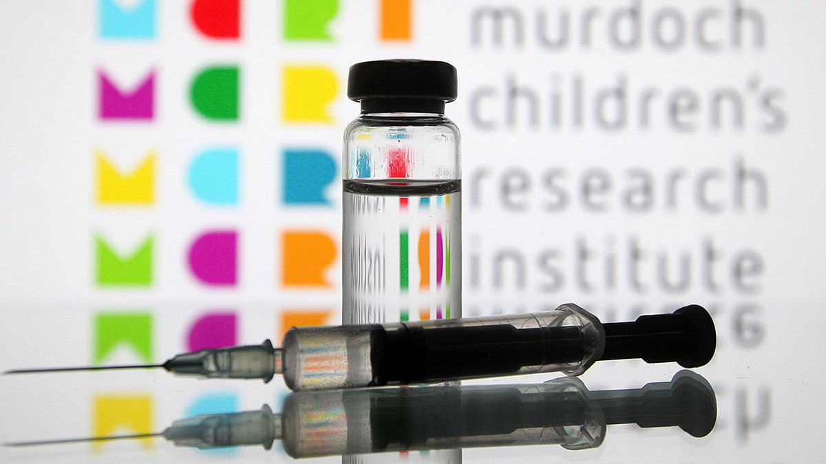 A medical syringe and a vial labeled COVID-19 vaccine are pictured in front of the Murdoch Children’s Research Institute logo.