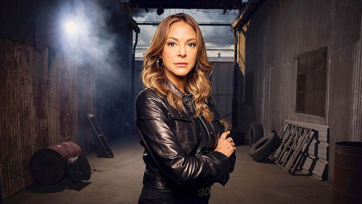 Eva LaRue with her arms crossed as she wears a leather jacket in a dark alley.