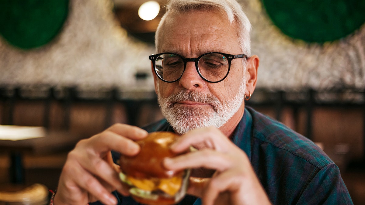 An old man with glasses and a beard and wearing an earring holds a burger in his hands at a restaurant.