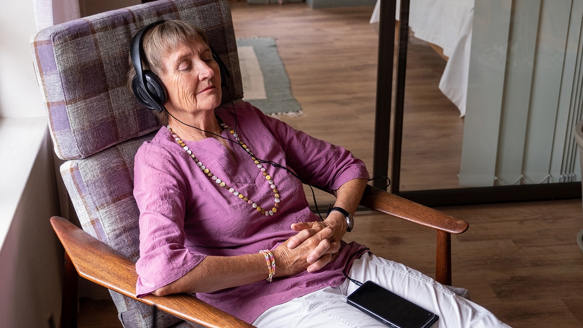 Senior woman relaxing in chair with eyes closed, hands folded listening to music with headphones on and smartphone on lap.