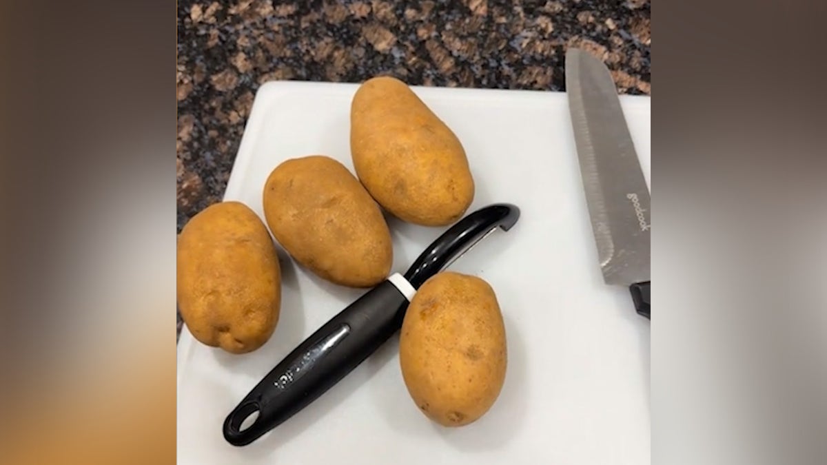 Potatoes on cutting board on counter with peeler and knife.