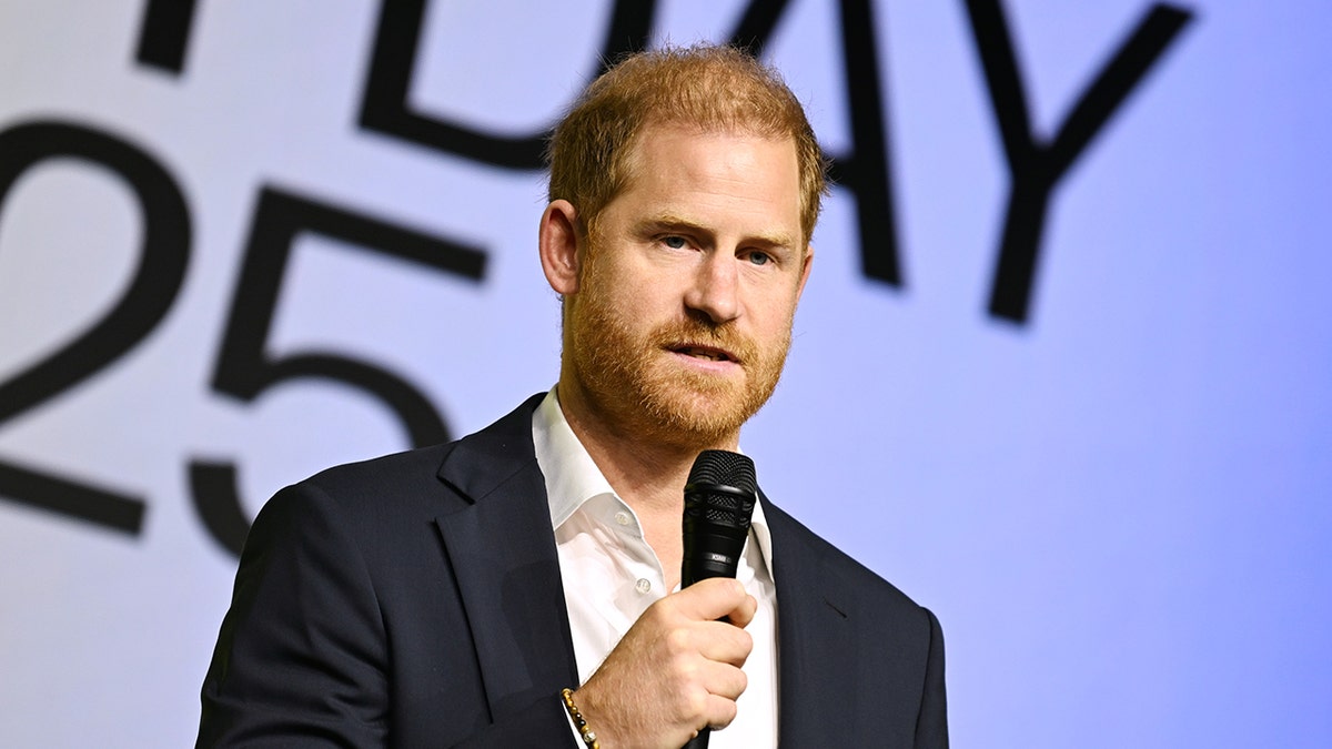 Prince Harry giving a serious speech wearing a dark blue blazer and a white shirt holding a mic.