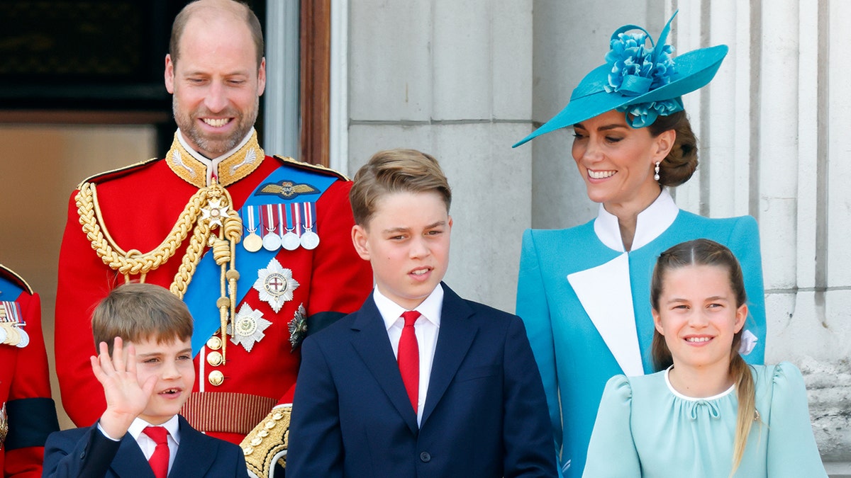 Prince William smiling in a red uniform on the Buckingham Palace balcony with his family.