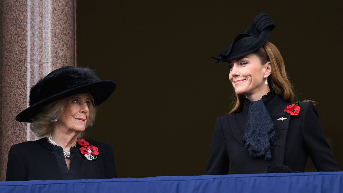 Queen Camilla and Kate Middleton smiling at each other on the balcony of Buckingham Palace.