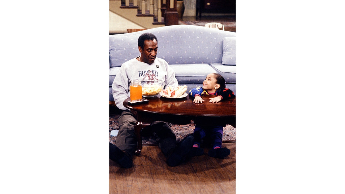 Bill Cosby and Raven Symone sit on the ground in a scene from the cosby show
