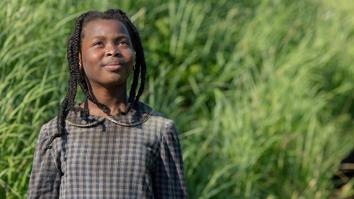 Main character in film "Sarah's Oil" in a checkered dress stands in a field of tall grass, looking upward with a hopeful expression.