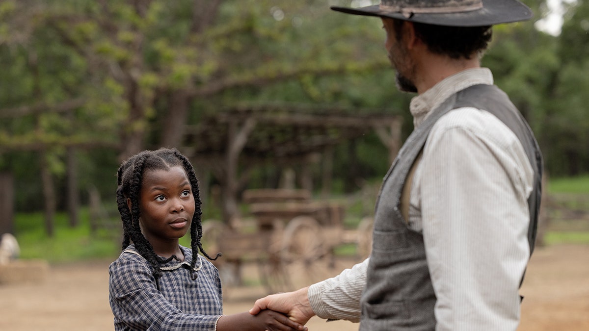 Zachary Levi's co-star shakes hands with a man wearing a hat and vest in a rustic outdoor setting.