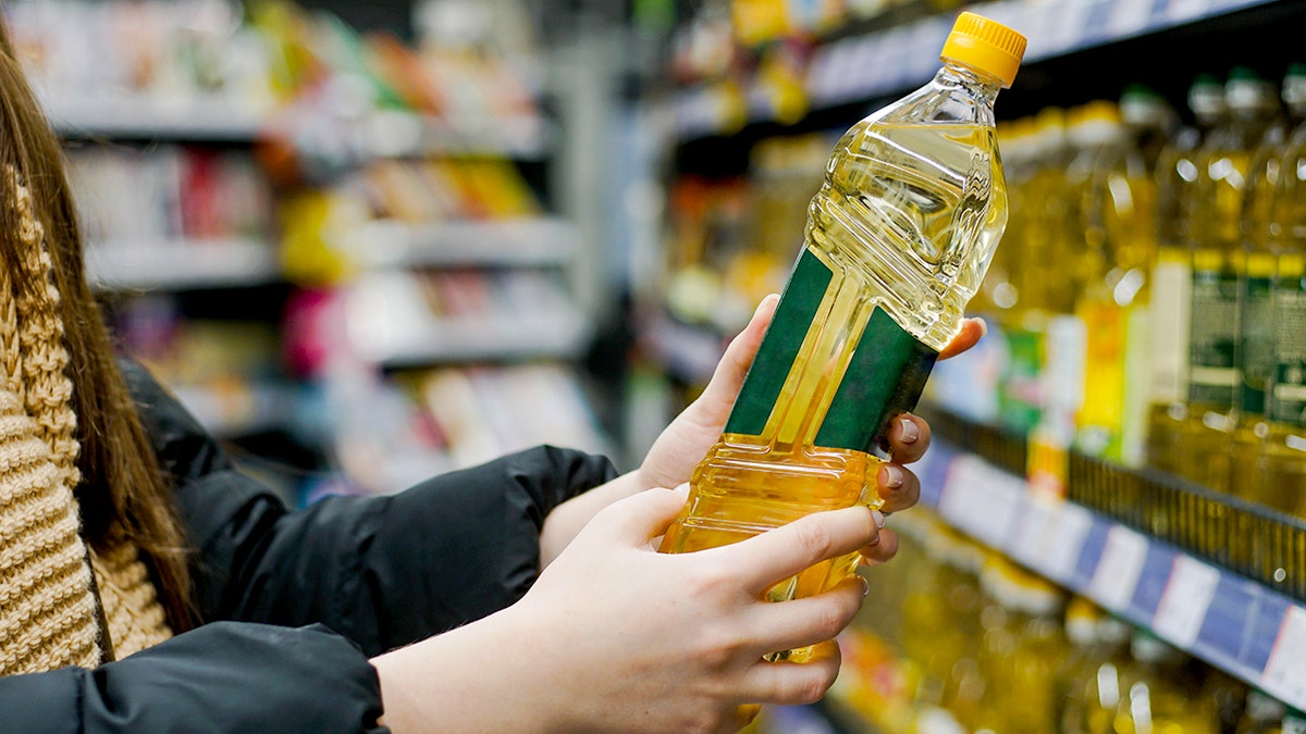 A close-up view of a woman's hand holding a bottle of sunflower oil at a supermarket.