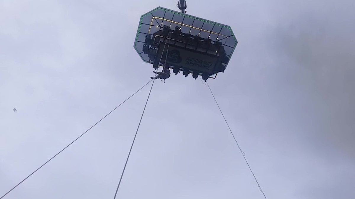 A view from the ground of a sky-dining attraction, where the firefighter is climbing a rope to rescue a family.