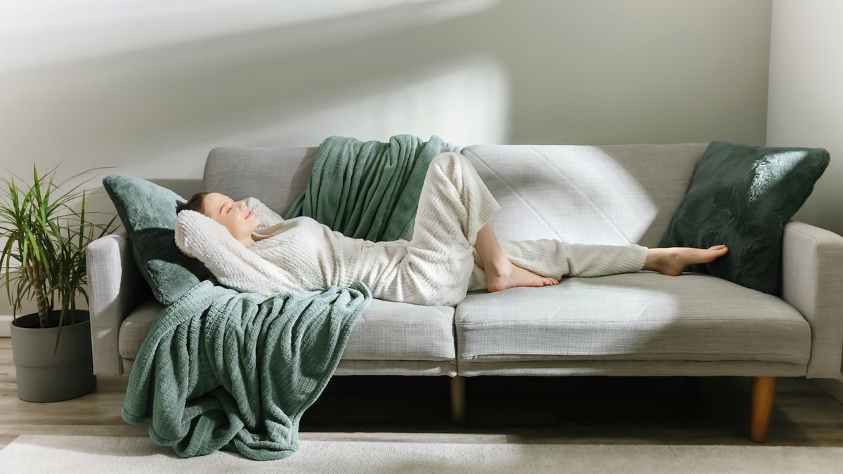 Woman in soft loungewear resting on a modern sofa with green blankets and pillows. 