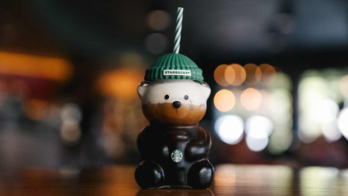 starbucks glass cup shaped like a bear with green beanie and striped straw filled with coffee drink
