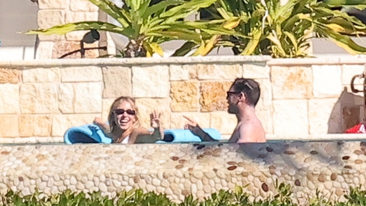 Sydney Sweeney is seen smiling and laughing while Scooter Braun gestures toward her as they relax in the pool, with stone walls and plants in the background at her Florida Keys home.