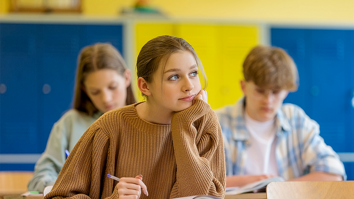 thoughtful female high school student sitting at the desk in the classroom and looking away with hand on chin.