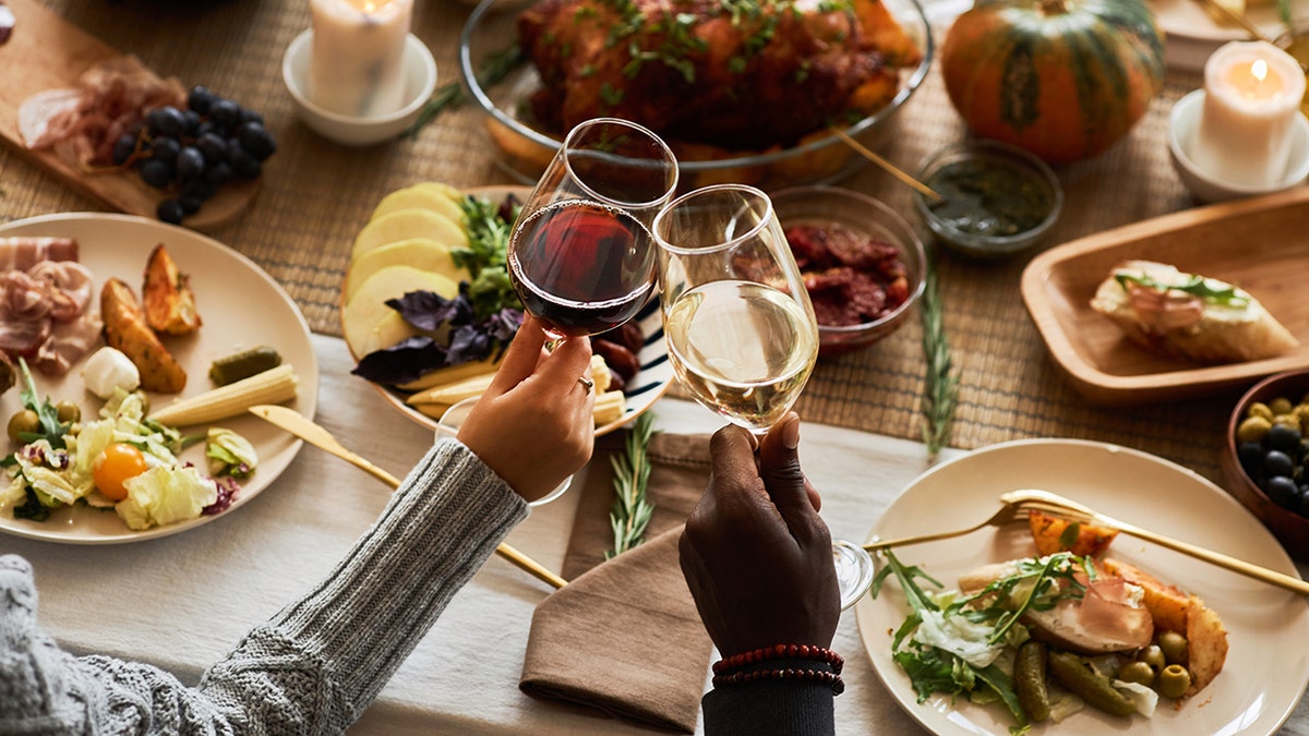 Two people toasting with red and white wine at Thanksgiving dinner, with food seen spread across table.