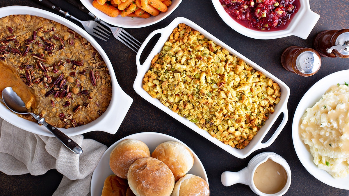 Spread of Thanksgiving sides laid out on table, including sweet potato casserole, stuffing, cranberry sauce, mashed potatoes, rolls, gravy and cranberry sauce.
