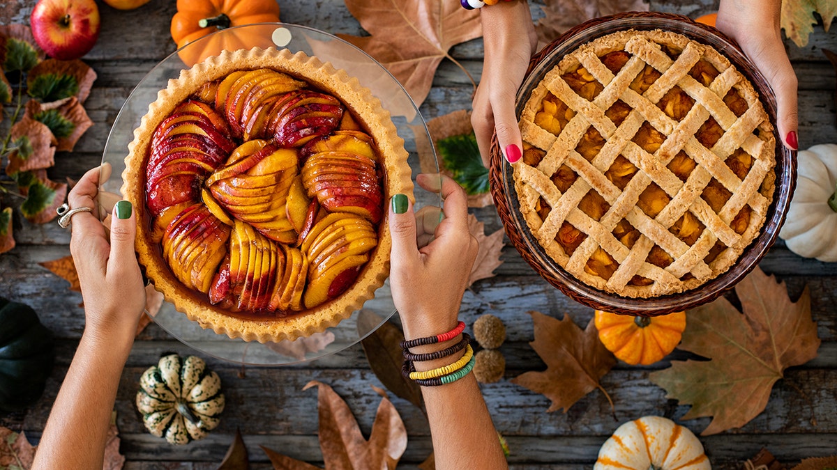 Hands holding out two different Thanksgiving pies, above table with pumpkins and other Thanksgiving decor.