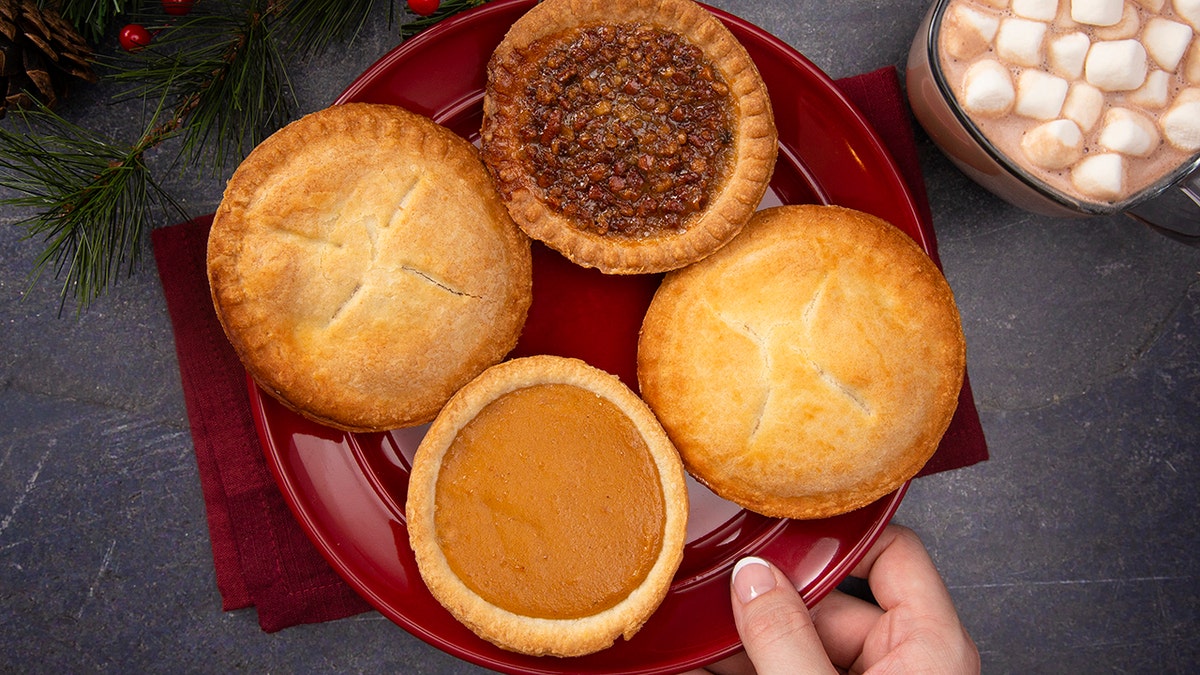 Hand holding plate of four different mini pies including pecan and pumpkin over a holiday table