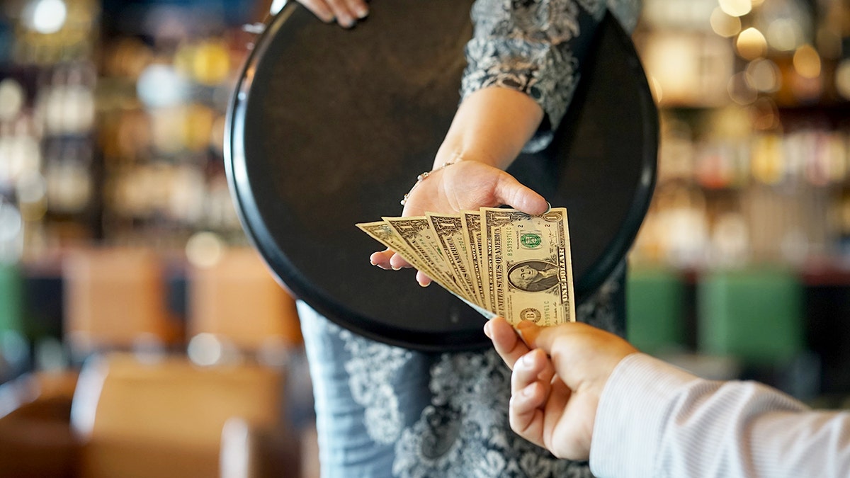 A man's hand holds five $1 bills to give to a waitress holding a carrying tray at a restaurant.