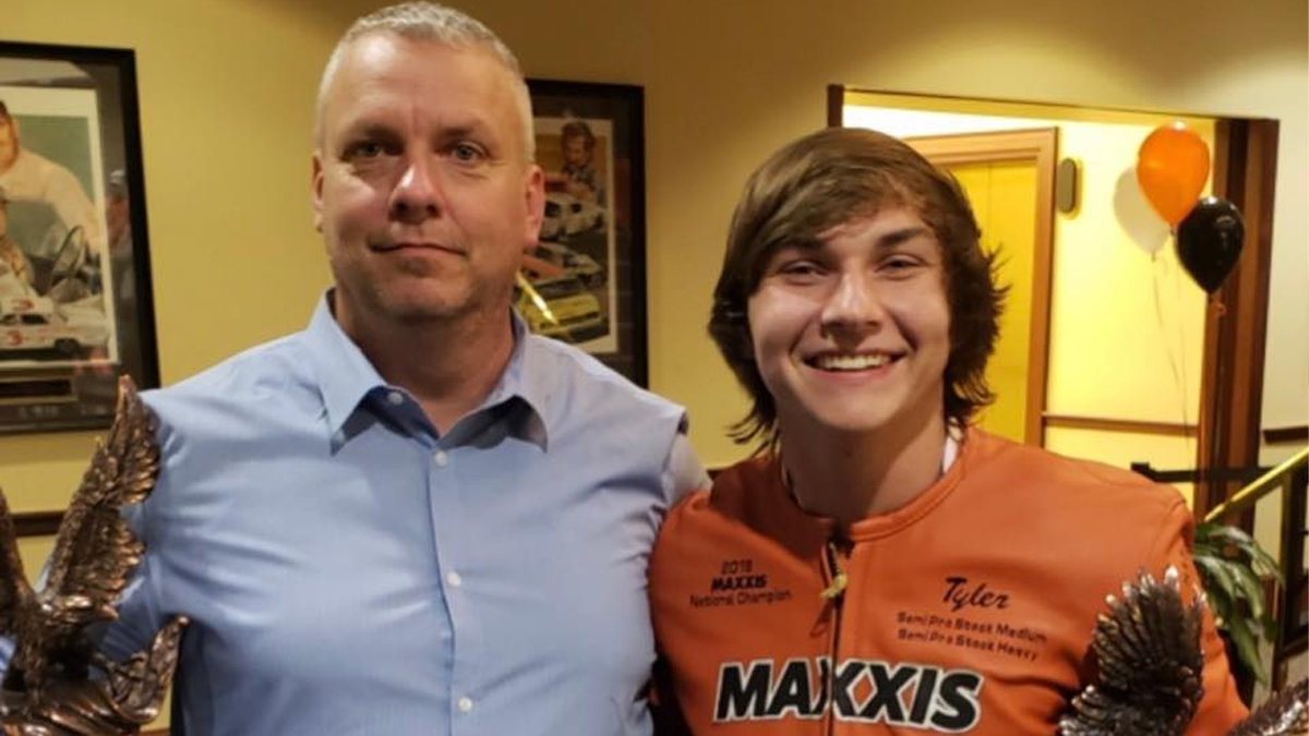 Tyler Weaver smiles wearing an orange racing jacket and holding a trophy next to his dad