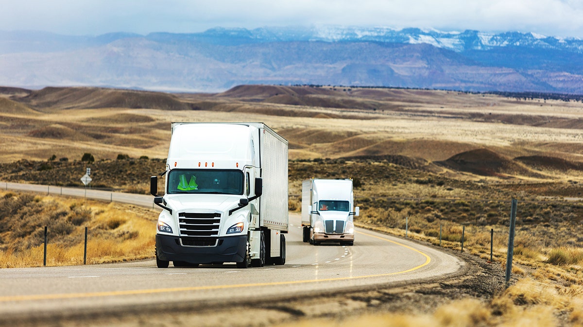 A pair of semi-trucks drive down a highway surrounded by mountains