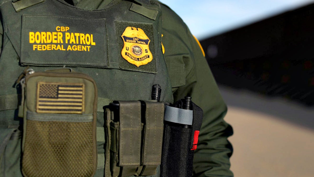 U.S. border patrol agent in green vest standing near border wall