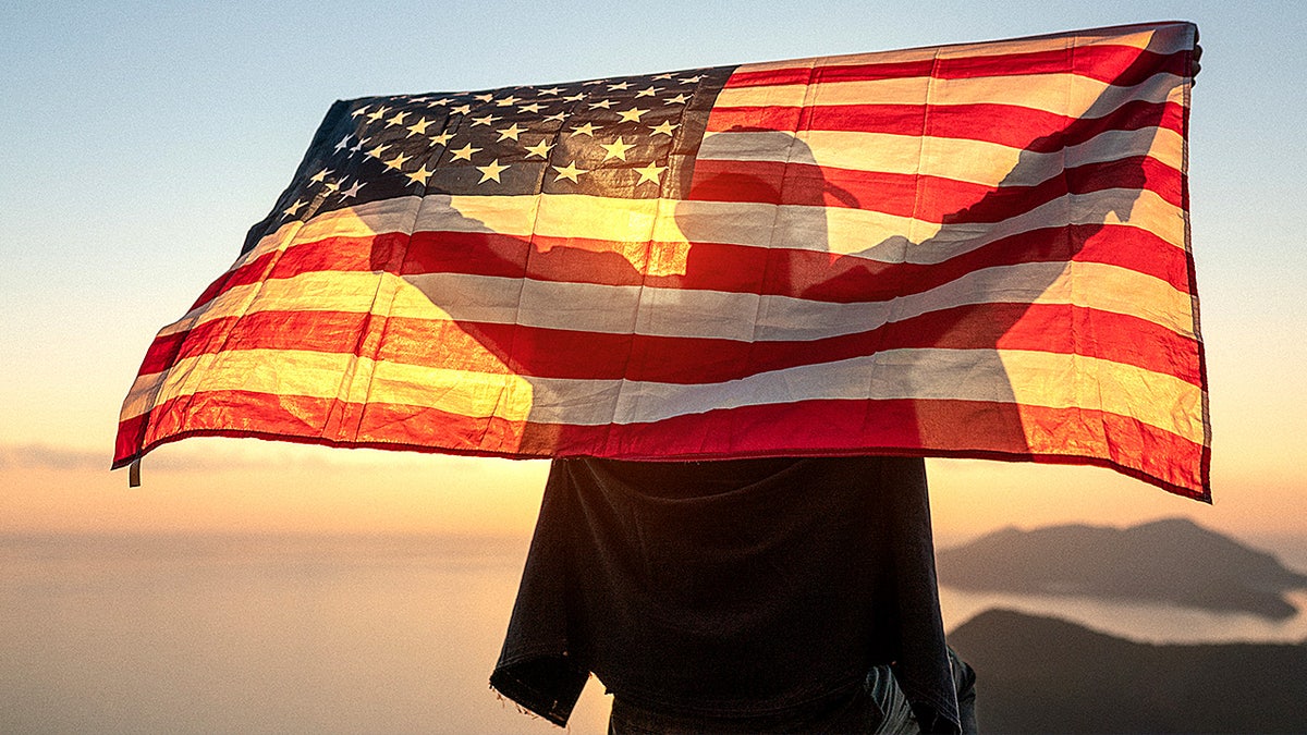A man holds an American flag above his head while looking toward the sun.
