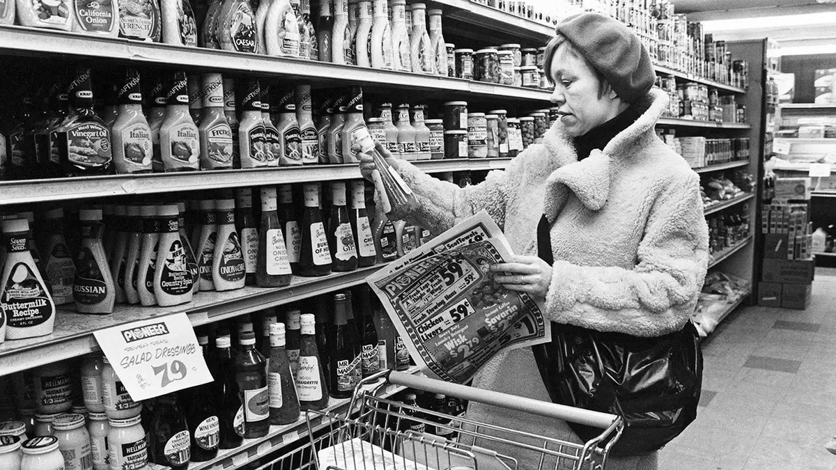 Black-and-white photo of a woman shopping in the Pioneer grocery store, New York City, USA, March 1983, looking at salad dressing in front of shelves of it, with hat on, purse in shopping cart in front of her.