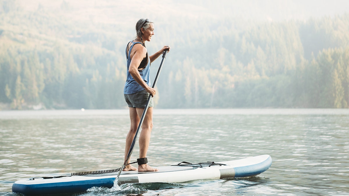 মহিলা একটি লেকের উপর একটি প্যাডেলবোর্ডের উপর দাঁড়িয়ে আছে