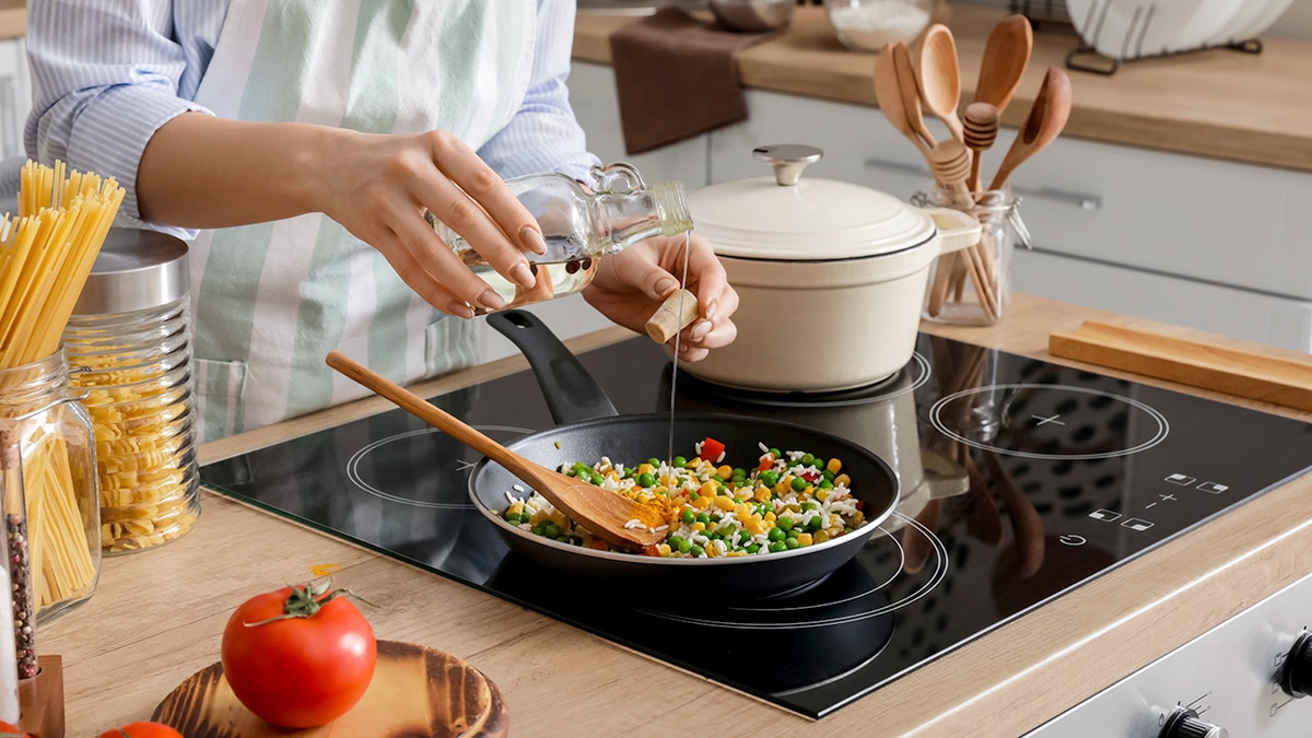 A woman pours olive oil from a glass over a stir-fry meal on a stove.
