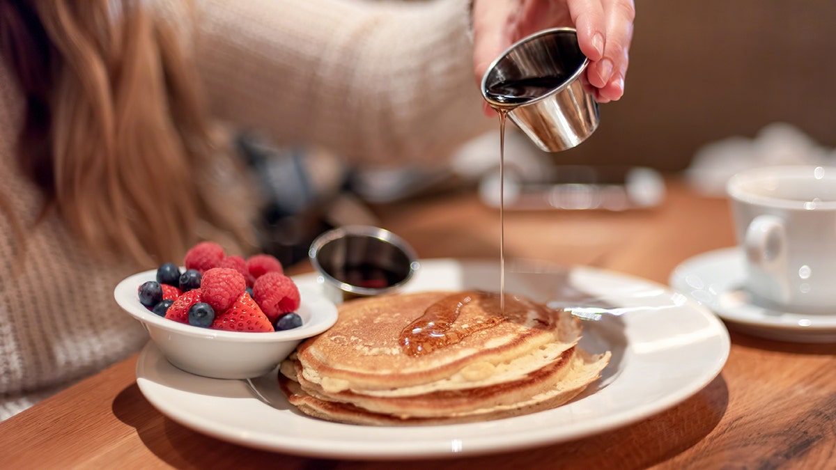 A woman is seen pouring syrup over a stack of pancakes with a side of blueberries, strawberries and raspberries.