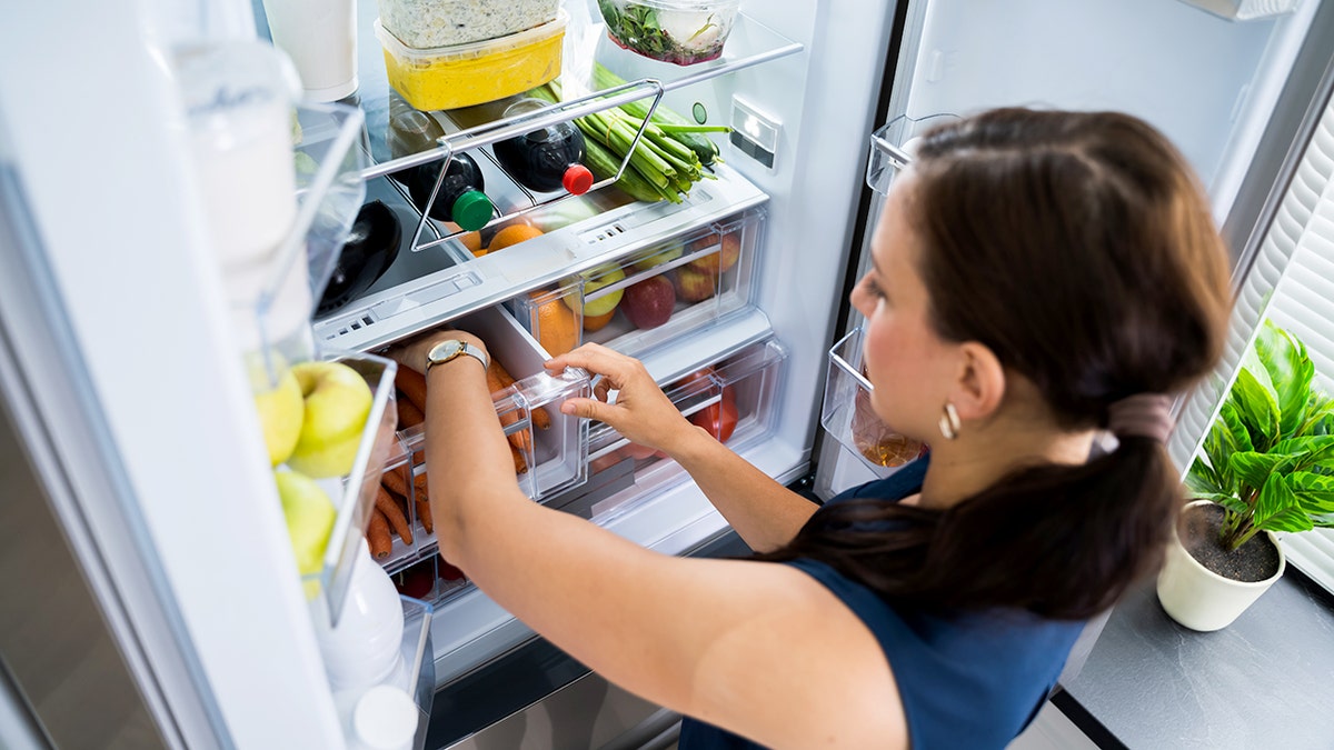 A woman wearing a watch reaches for a carrot inside the freezer compartment of the refrigerator.