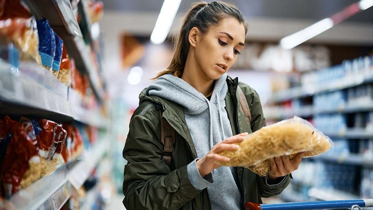 A young woman looks at a bag of pasta in grocery store, seen with cart in front of her.