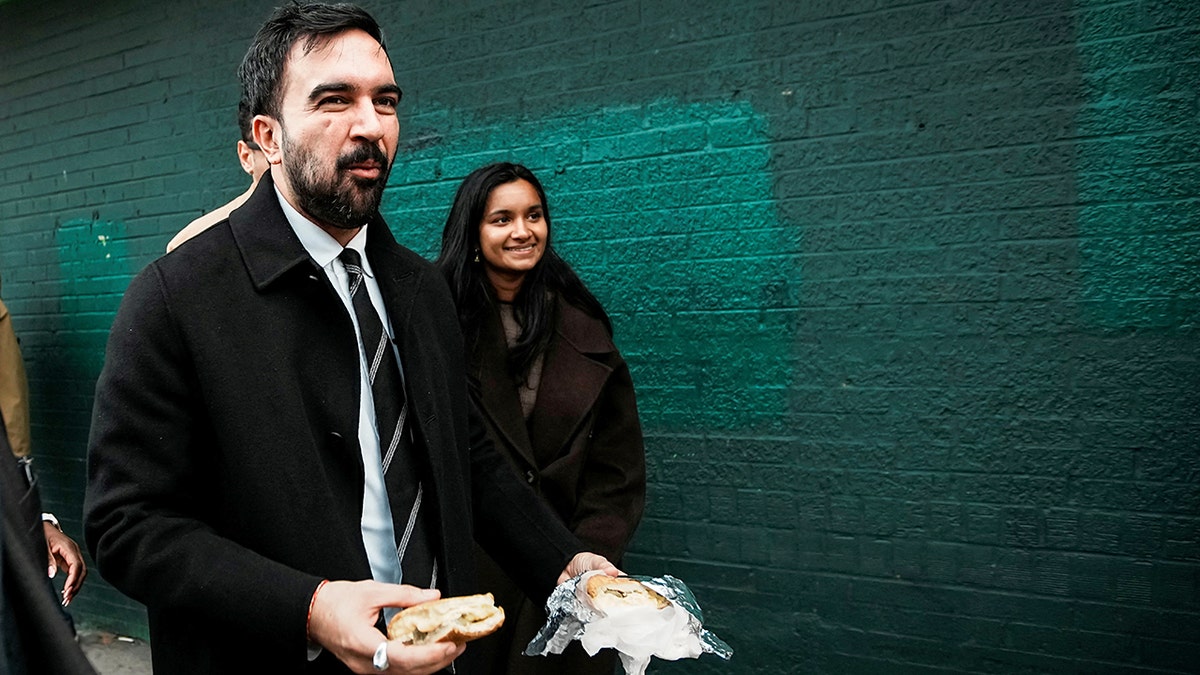 Zohran Mamdani eats a sandwich as he walks past a green brick wall.