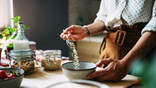 woman in kitchen pours dried oats into a bowl from a spoon