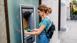 A woman wearing a teal shirt and jeans puts her card into an ATM.