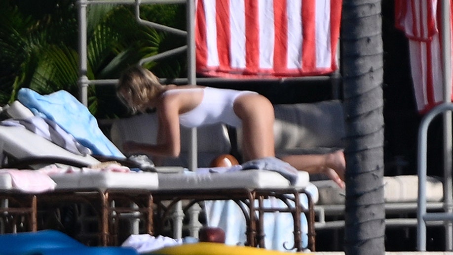 Actress Sydney Sweeney is seen kneeling on an outdoor lounger in a white swimsuit, organizing towels and belongings during a sunny day by the pool.