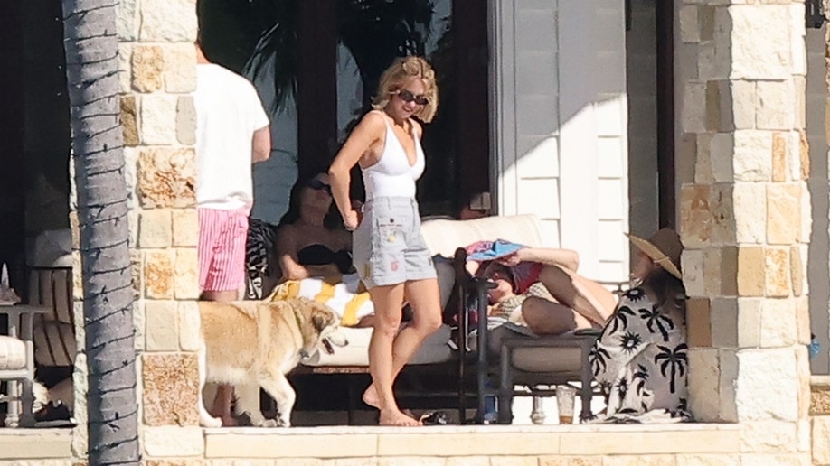 Actress Sydney Sweeney, wearing a white swimsuit and gray shorts, is seen standing on the patio beside friends and a dog at her Florida Keys property.