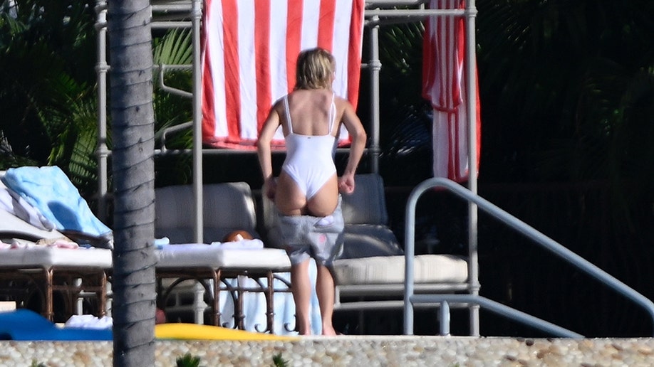 Actress Sydney Sweeney is photographed from behind in a white swimsuit as she pulls on her gray shorts near lounge chairs and striped towels at her Florida Keys property.