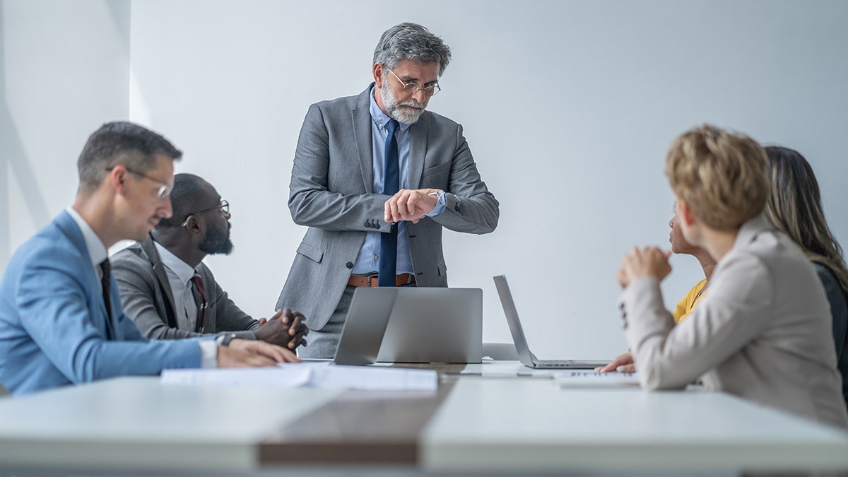 A group of employees are sitting in a conference room, and the manager is checking the time on his watch.