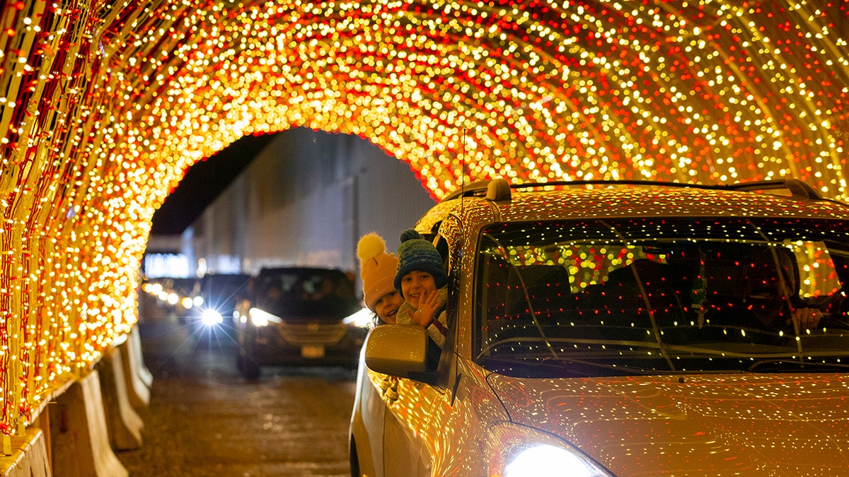 Children smiling and waving from a car driving through a tunnel of golden holiday lights at a festive drive-through light display.