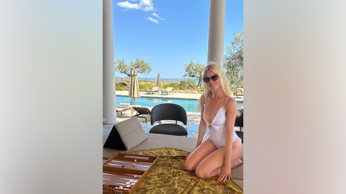 Claudia Schiffer kneels beside a pool wearing a white one-piece swimsuit and sunglasses, smiling during her Aug. 25, 2025, Instagram birthday photo with lounge chairs and greenery in the background.