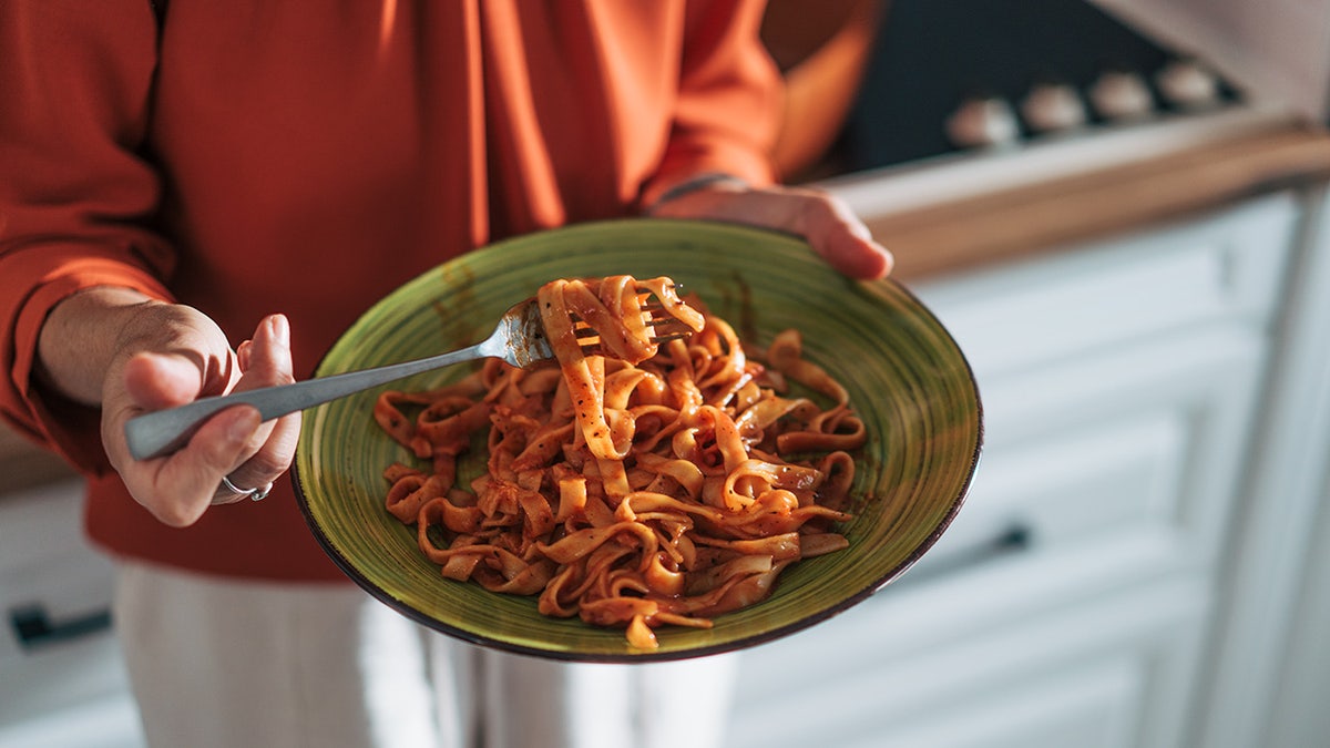 Older woman wearing sweater holding plate of pasta with tomato sauce in kitchen.
