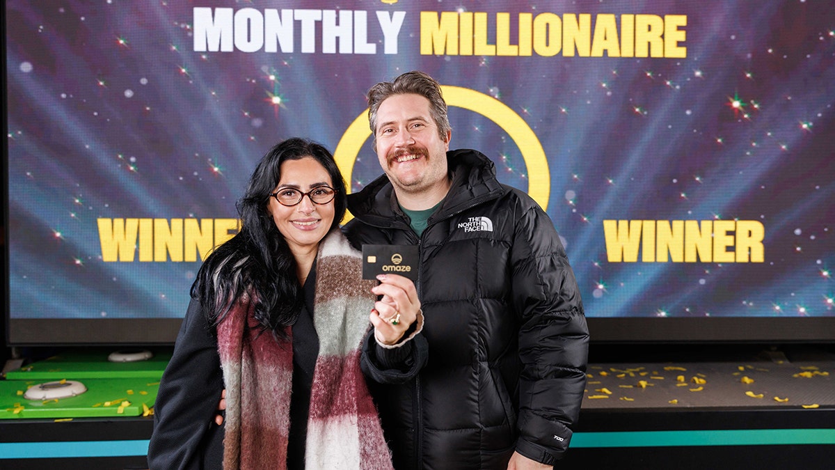 Two smiling people hold a black Omaze card in front of a 